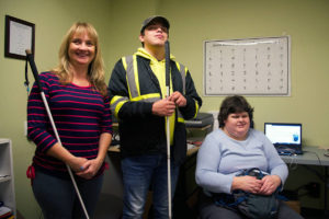 Elizabeth Lalonde (left) executive director at the Pacific Training Centre for the Blind, Alex Jurgensen and Heidi Propp each help blind and partially-sighted people learn how to navigate the internet. However, some frequent shortcomings make accessing many websites very difficult. (Nicole Crescenzi/News Staff) [Alt Text: A woman in a red and blue striped shirt using a white cane stands next to a young man in black and yellow, also using a white cane. Seated on the right, a woman in blue sits next to a computer. On the wall in the background is a Braille chart.] Photo: Victoria News/ Black Press Media